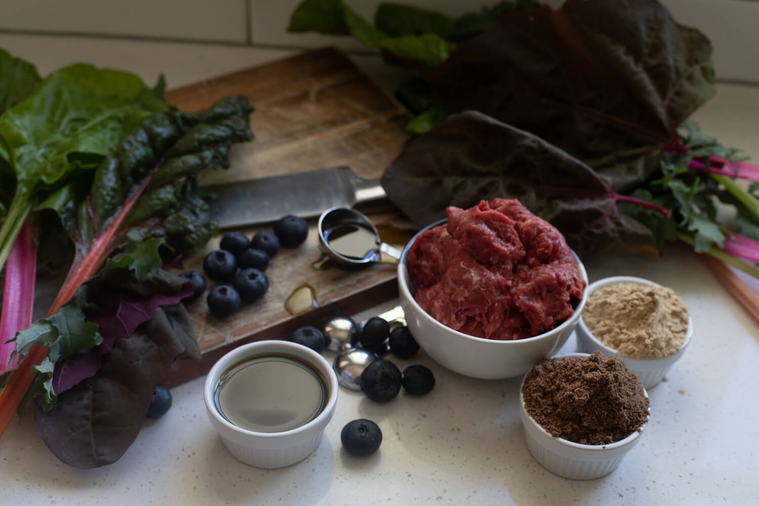 The ingredients for the Huntaway wild raw venison dog food sitting on a white kitchen counter including: a bowl of venison meat and offal; a handful of blueberries; two clusters of silverbeet leaves; 3 ramekins filled with flaxseed oil, farmers yeast and chicory root; a tablespoon of apple cider vinegar; a chopping board and a kitchen knife